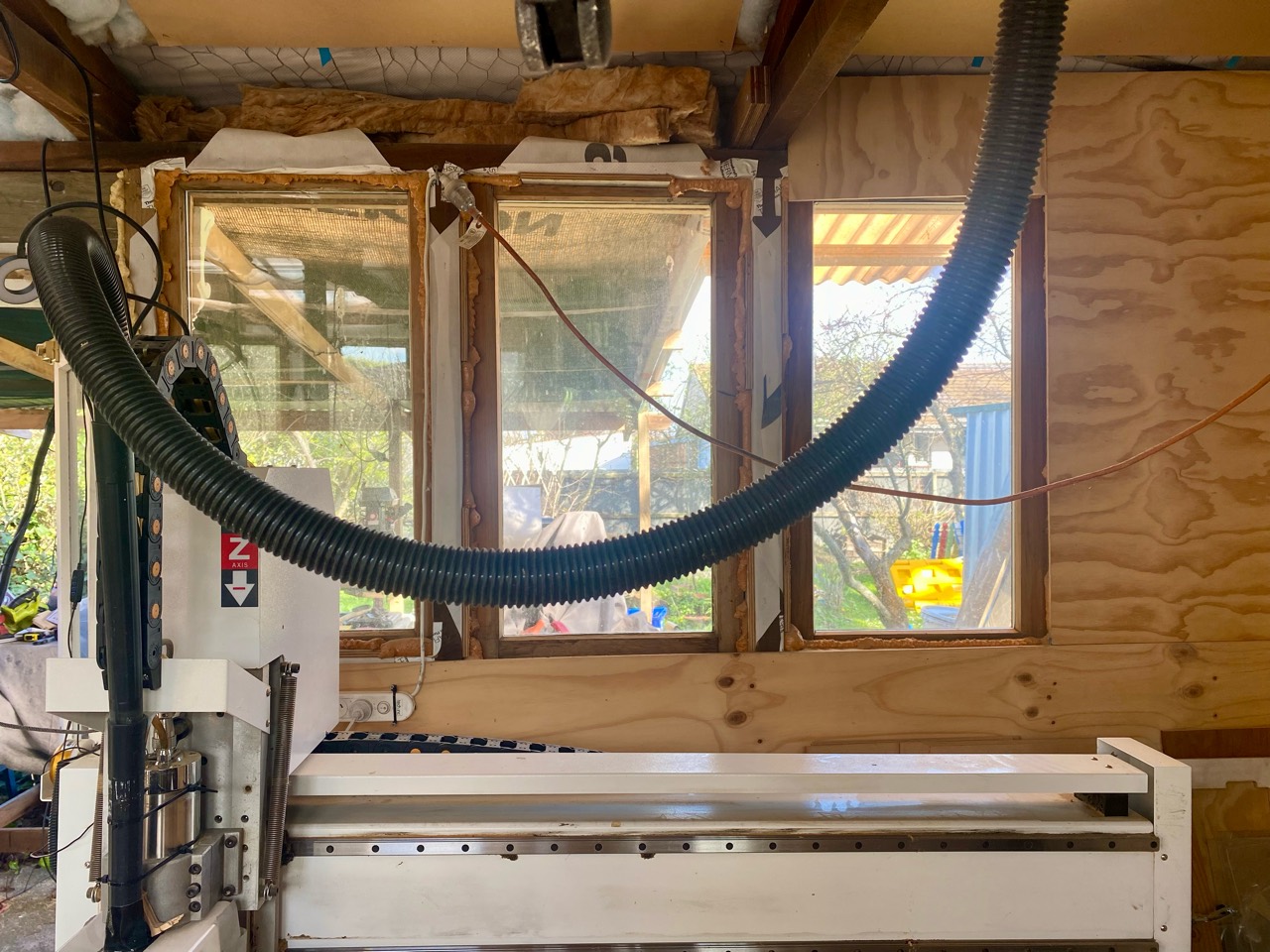 Wide view of the CNC router, dust hose, timber walls, and garden beyond.